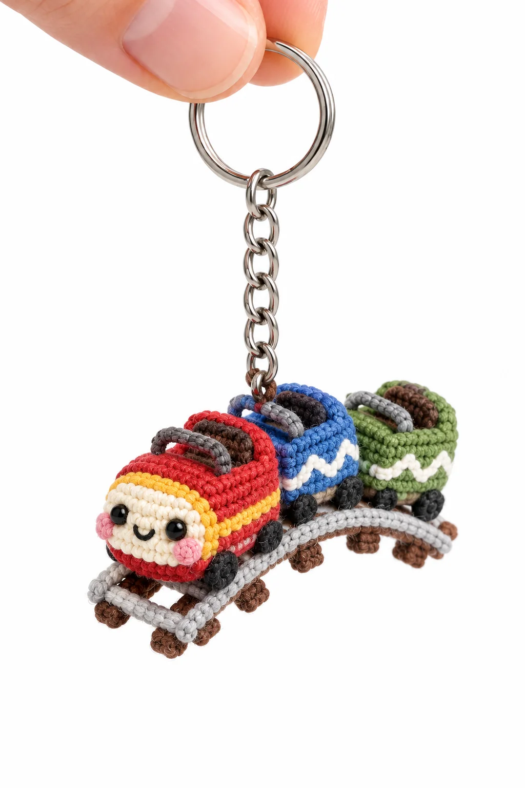 Colorful crochet train keychain with a smiling red engine pulling blue and green carriages on a grey track with wooden sleepers